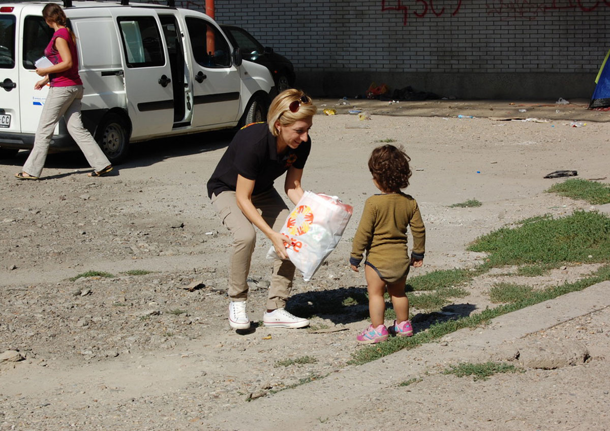A woman wearing a navy top bends down to offer a bag to a small child. The bag has the CARE logo on it.
