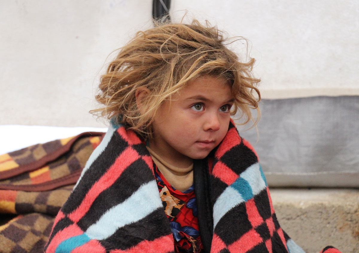 A young child affected by the Syrian refugee crisis sits in a tent with a striped blanket wrapped around them. Refugees in Syria struggle to access basic needs like food, shelter, and healthcare.