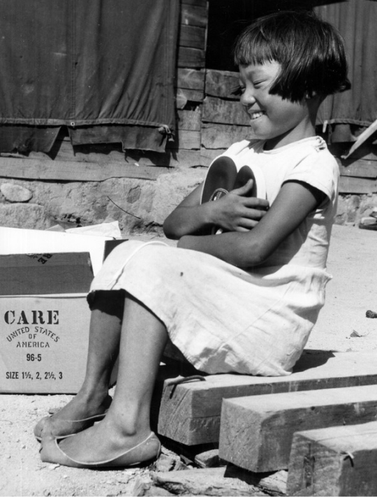 A young Korean girl sits on a curb and smiles while hugging a pair of shoes.