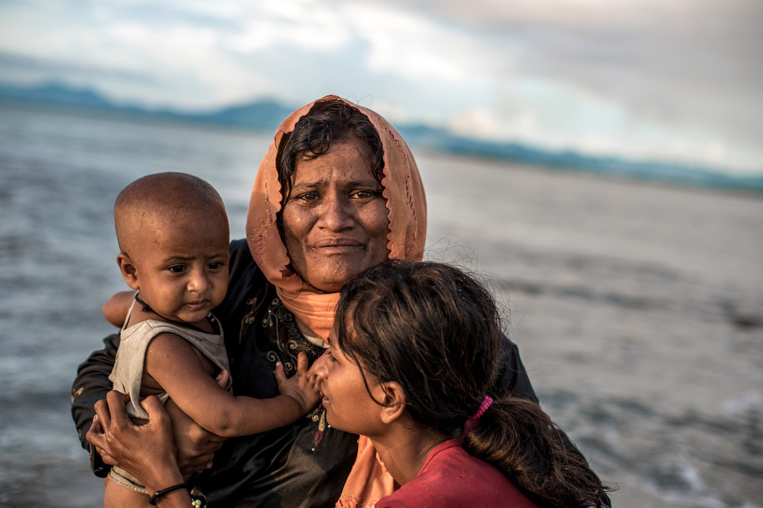A distressed woman huddles with two children, one of whom is a baby, in front of the ocean.