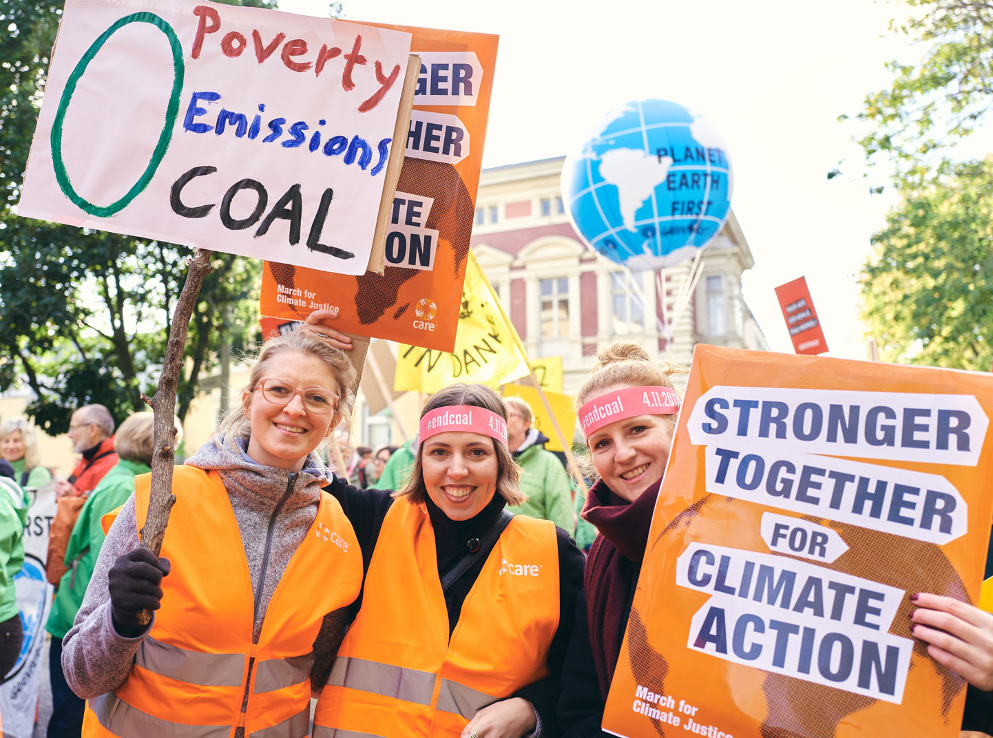 Three women wearing orange vests smile at a CARE for Climate Action march. One is holding a sign that says, 