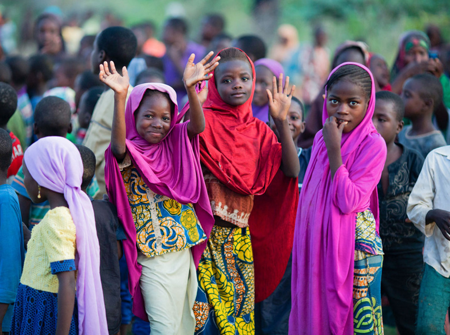 A group of children gather in the Sahel region in Niger. Three of them are smiling and waving at the camera.