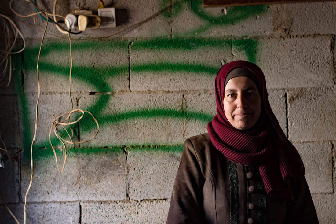 A woman wearing a burgundy head scarf stands in front of a concrete wall marked with green spray paint.