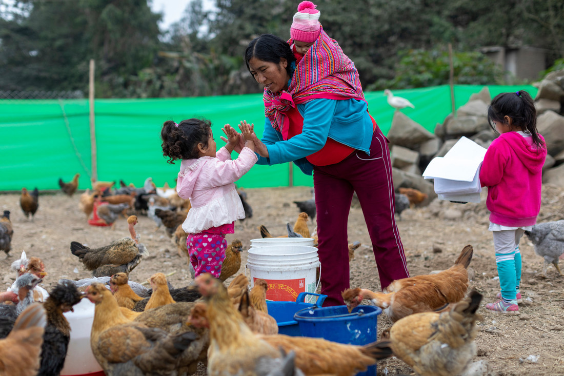 A woman carrying a baby on her shoulders high fives a young girl with both hands. They're surrounded by chickens eating on a farm.