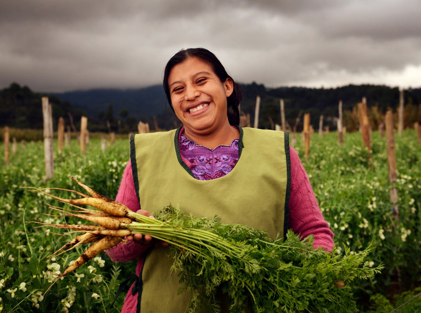 A woman stands in the middle of a large field and beams while showing off some carrots she's just pulled from her crop garden.