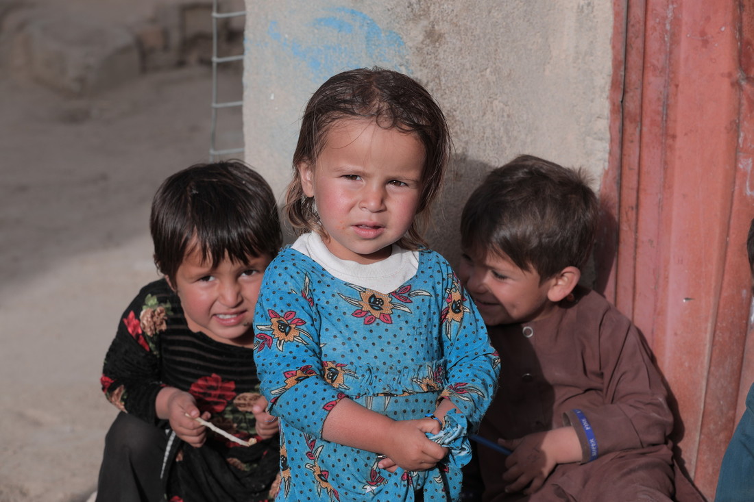 Three Afghan children sit together and lean against a house.